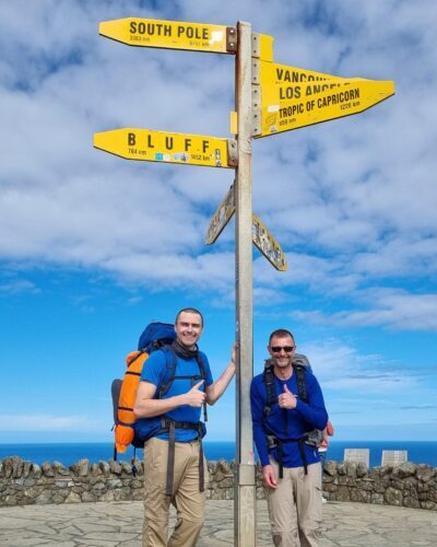 Two men standing under signpost at Cape Reinga. Both are wearing rucksacks.