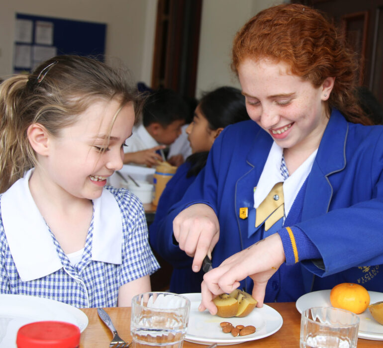 Older school girl helping junior student at the lunch table by cutting up fruit for her.