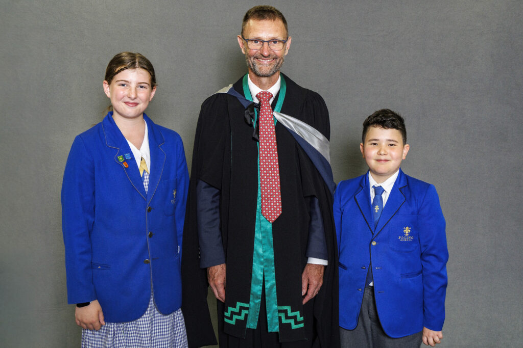 Man wearing academic gown standing with two students in blue blazers smiling for the camera.